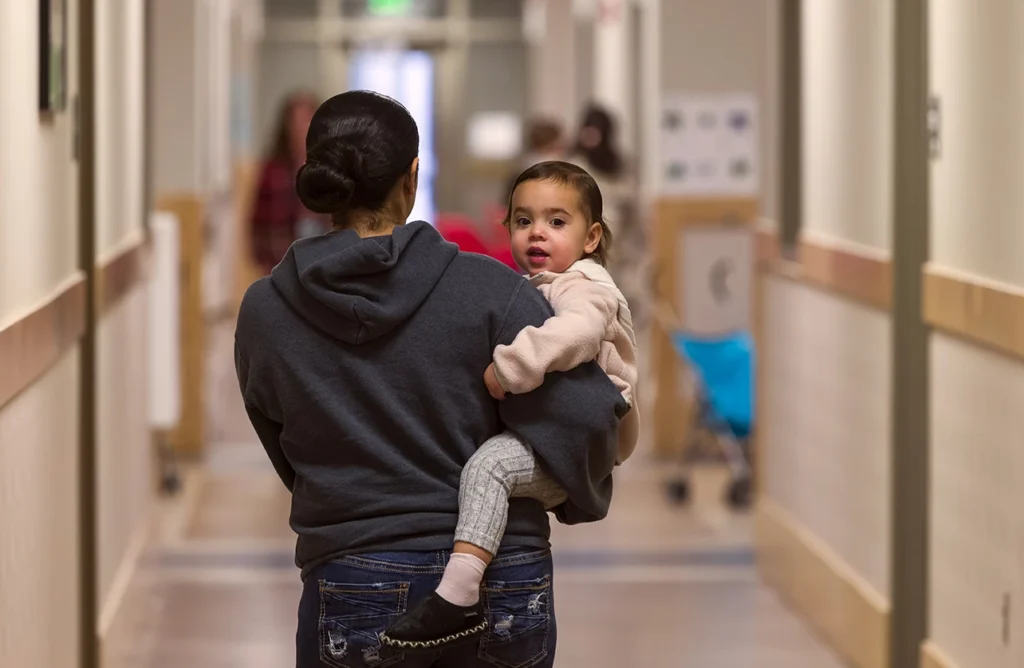A caregiver carries a toddler down a clinic hallway, the child looking back at the camera