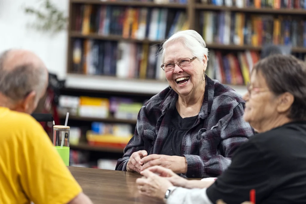 An older woman laughs joyfully while gathered with peers around a table in a community space lined with bookshelves