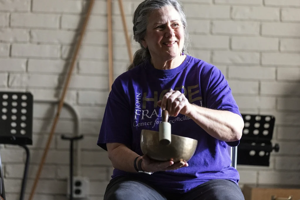 A smiling woman in a purple John Hope Franklin Center t-shirt plays a Tibetan singing bowl while seated