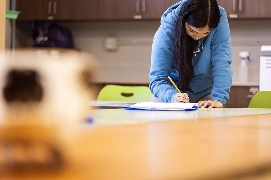 A young woman in a blue hoodie writes on paper at a desk in a classroom or study space