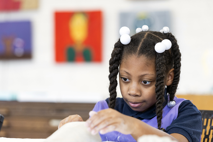 A young girl with braided hair and white beads concentrates while working with clay in an art classroom