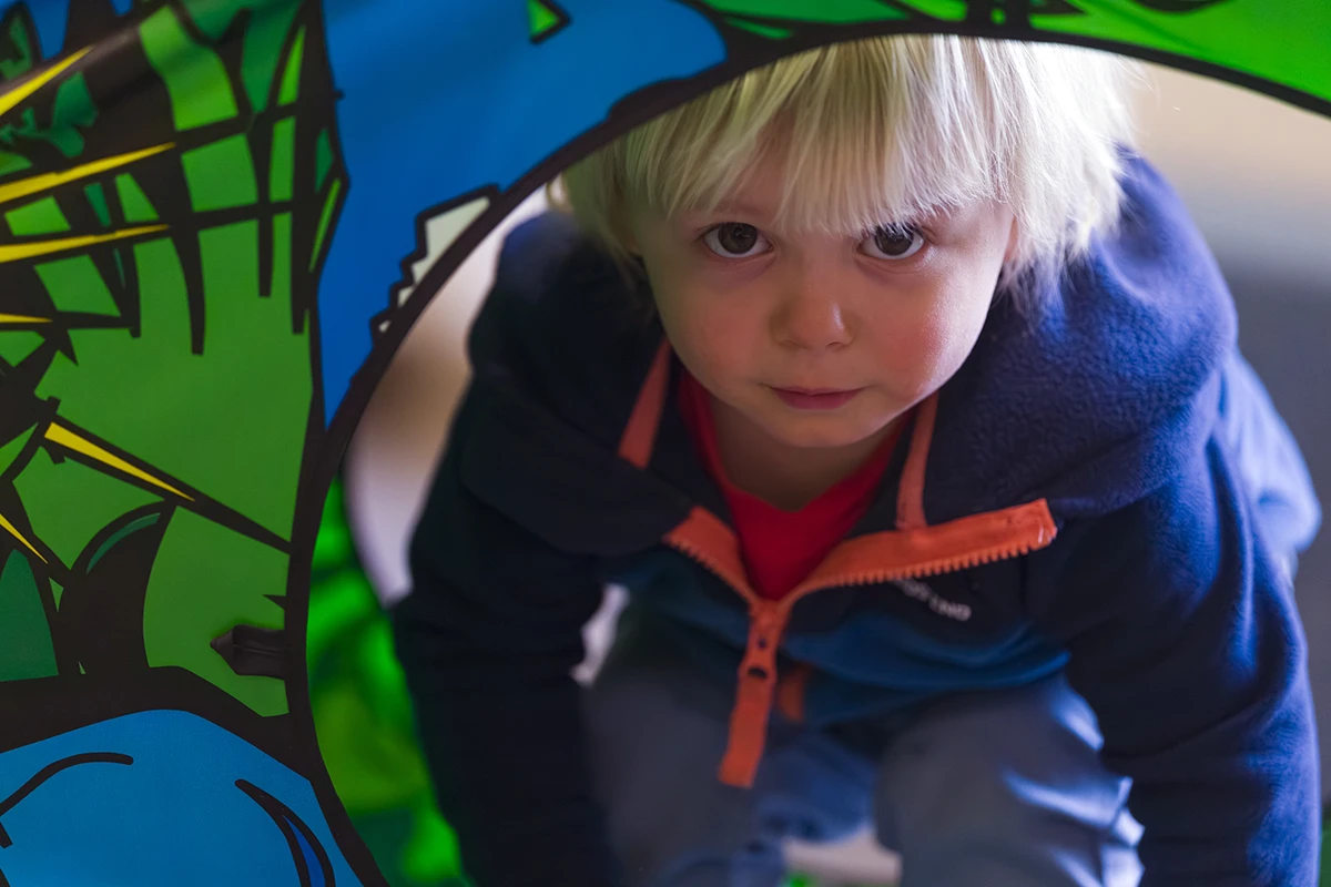 Young blonde child peering through a colorful play tunnel with a curious expression, representing youth development