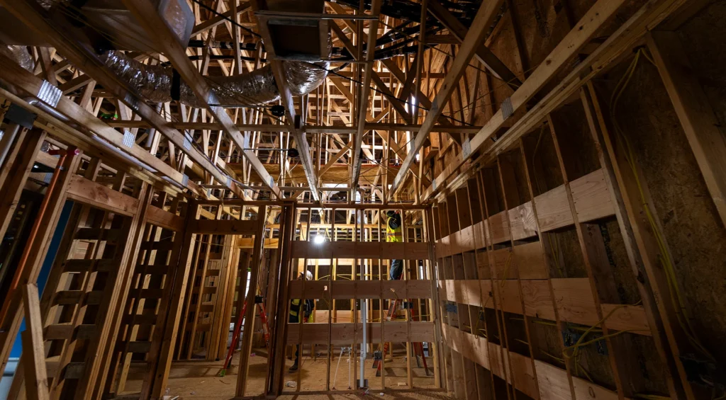 Interior wood framing of an affordable housing construction site with workers in safety vests visible among exposed beams