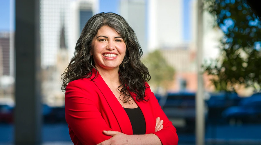 Woman in red blazer smiling with arms crossed against blurred Tulsa city skyline backdrop