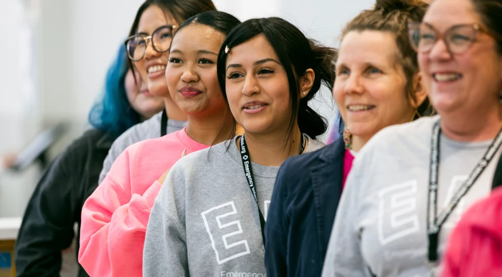 A diverse group of smiling women wearing Emergency branded sweatshirts stand together in a classroom setting