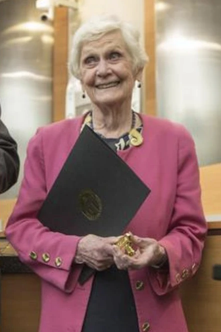 Elderly woman in pink blazer smiling while holding a black folio and small gold award at a formal recognition ceremony