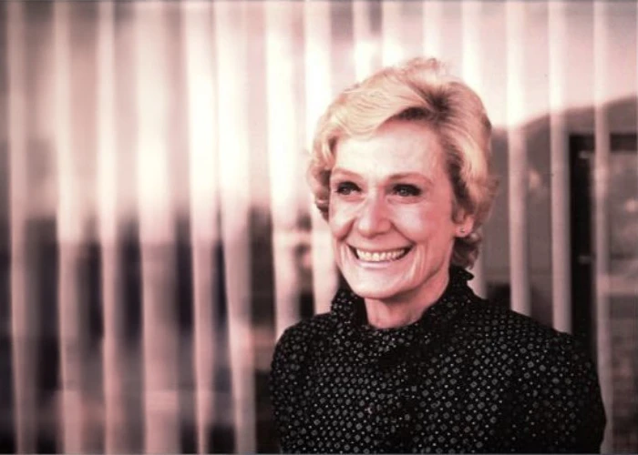 Smiling older woman with short blonde hair wearing a dark patterned blouse standing before vertical curtains
