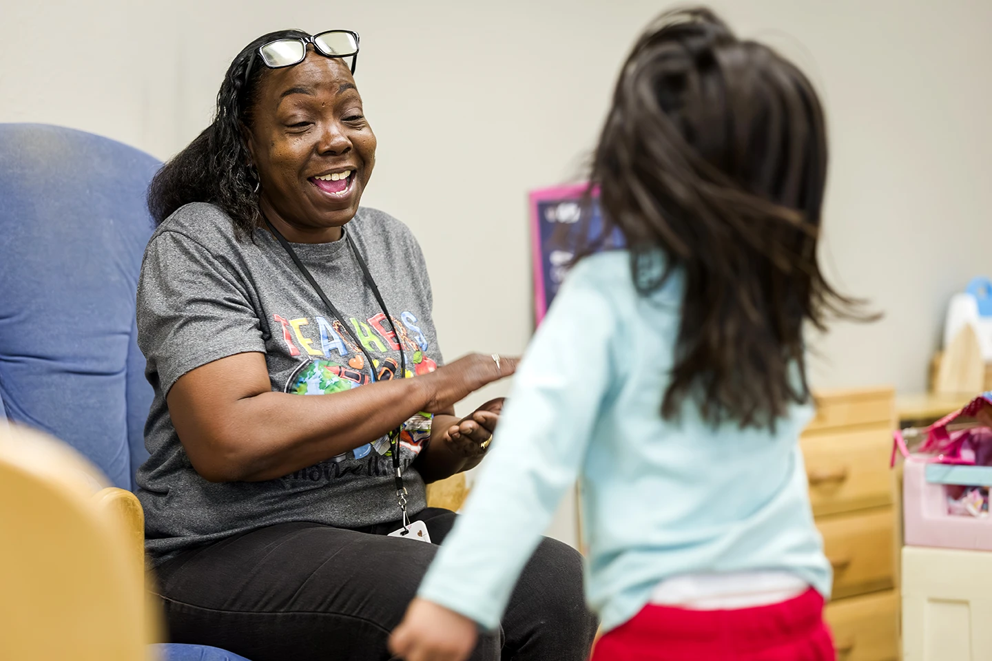 A smiling teacher holds a young girl's hands during a joyful interaction in a classroom setting