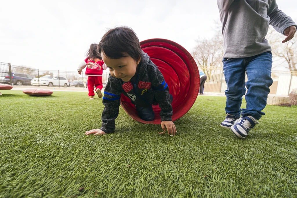 A young child crawls out of a red play tunnel on artificial turf at an outdoor playground
