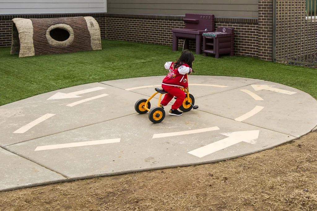A young child in a red outfit rides a tricycle on a circular concrete path with directional arrows at a playground