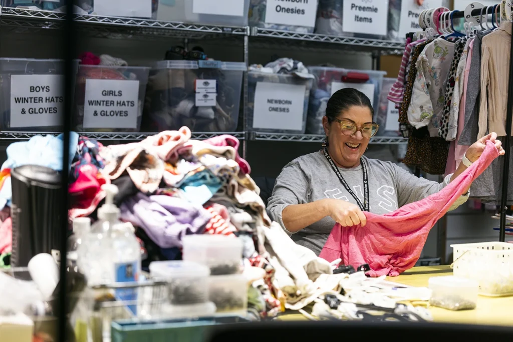 A smiling volunteer sorts donated children's clothing at an organized relief organization with labeled storage bins