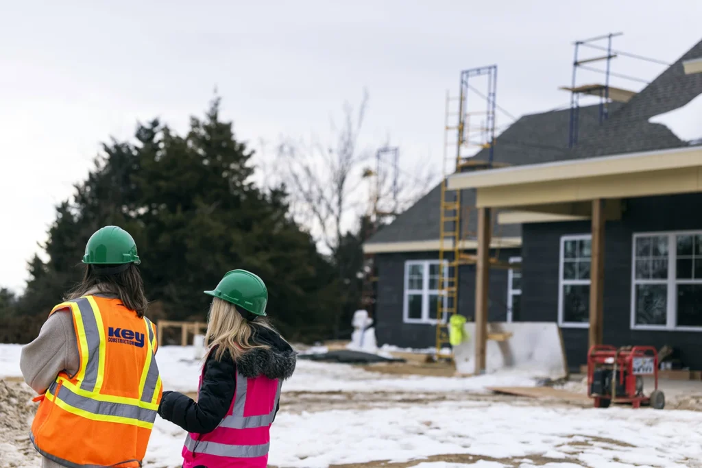 Two people in hard hats and safety vests survey a home under construction in winter
