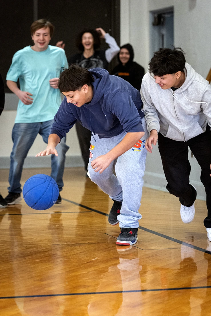 Teens playing basketball in a gym, laughing and chasing a blue ball across the court
