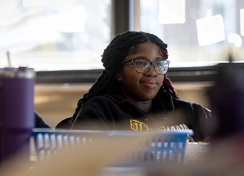 Young woman with braided hair and glasses smiles while seated at a table in a casual indoor setting