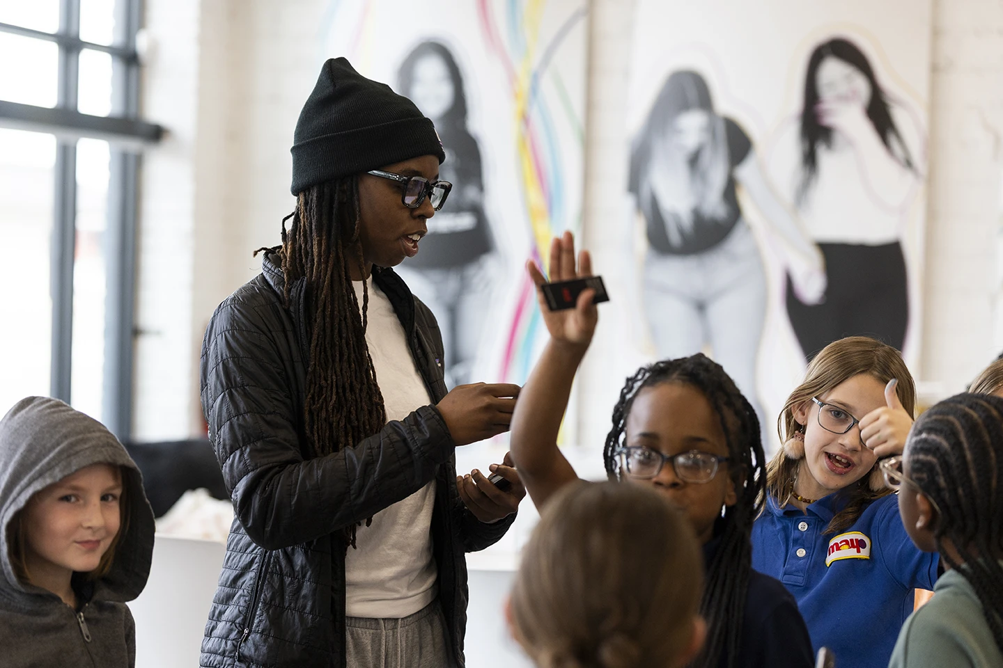 A young adult mentor engages a group of elementary school children in a discussion at an art-filled community space