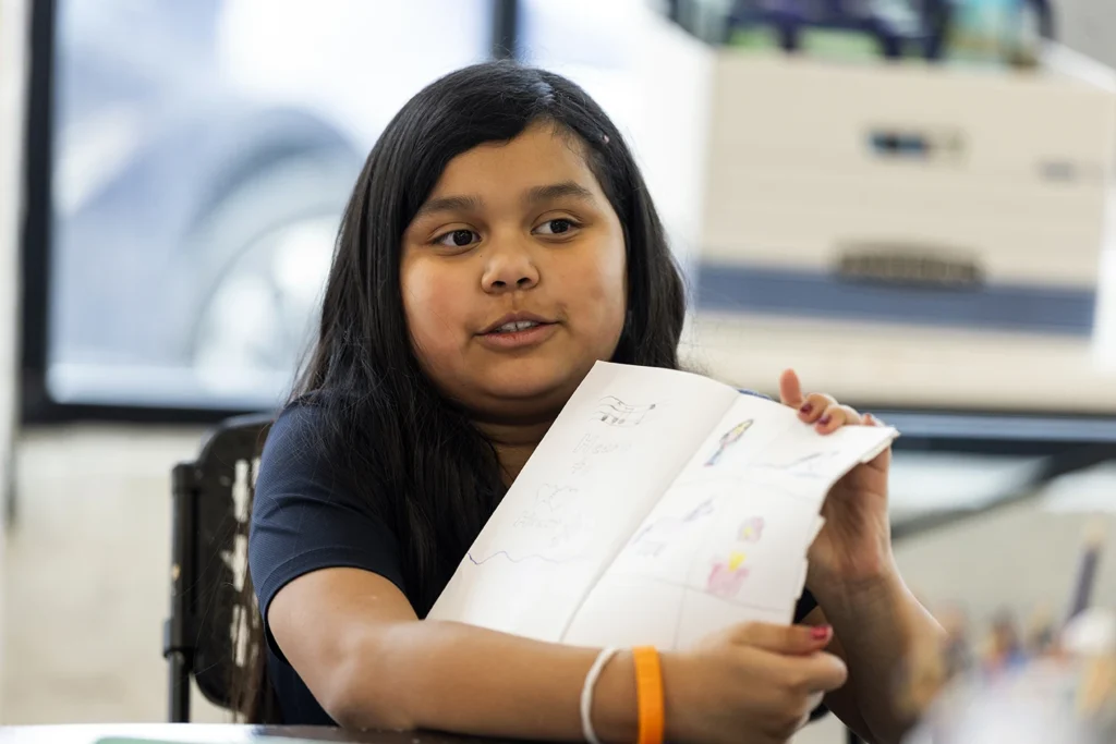 A young girl proudly holds up her hand-drawn artwork at her desk in a classroom