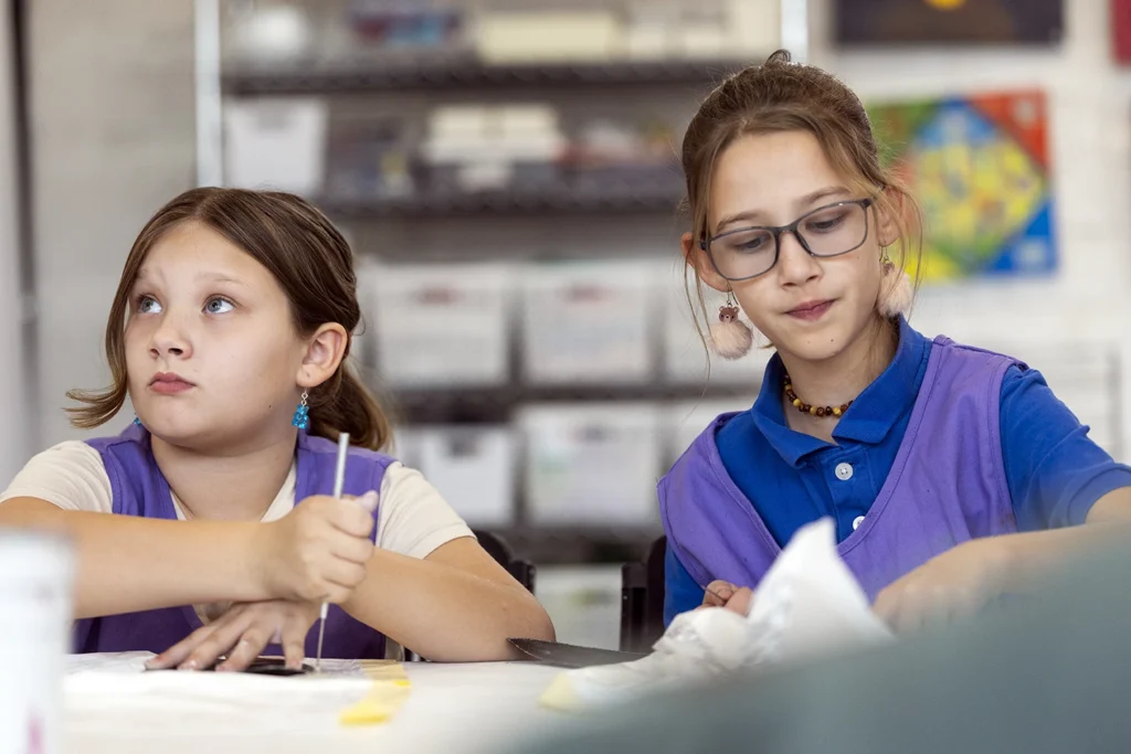 Two young girls in matching purple vests work together at a classroom desk during a school activity