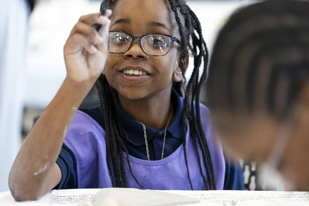 A smiling young girl with braids and glasses raises her hand eagerly during a classroom activity in Tulsa