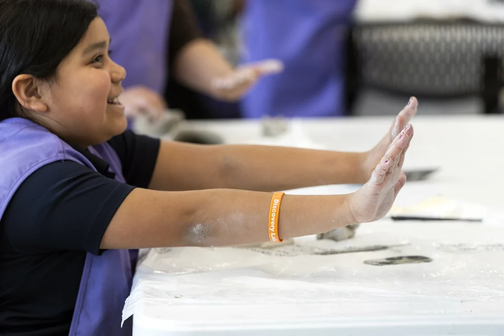A smiling child holds up clay-covered hands during a hands-on Discovery Lab activity while wearing a purple apron