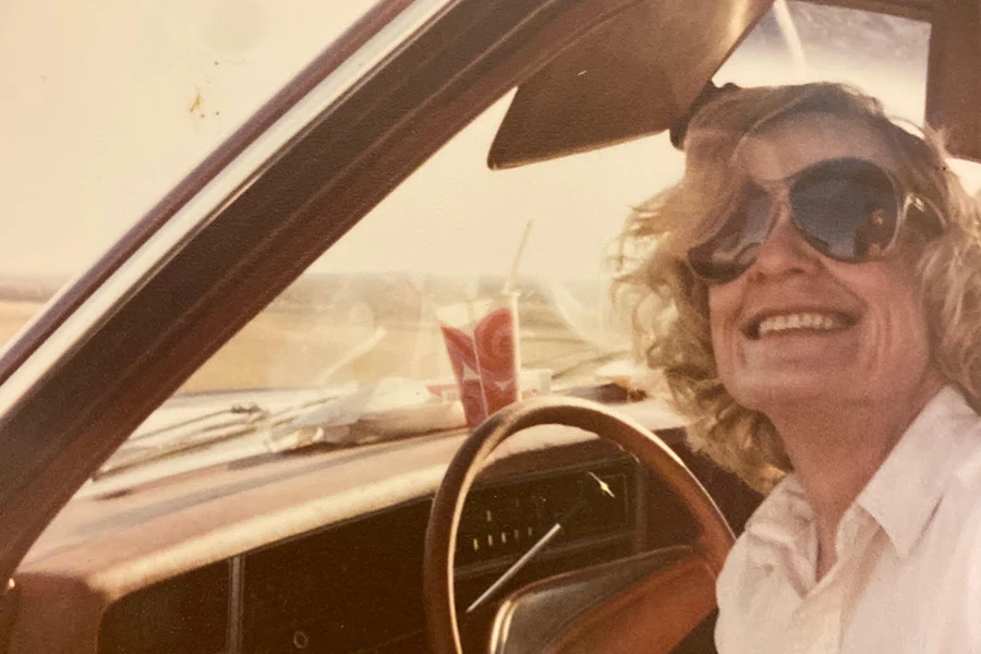 Smiling woman with curly blonde hair and sunglasses driving a vintage car on a sunny day