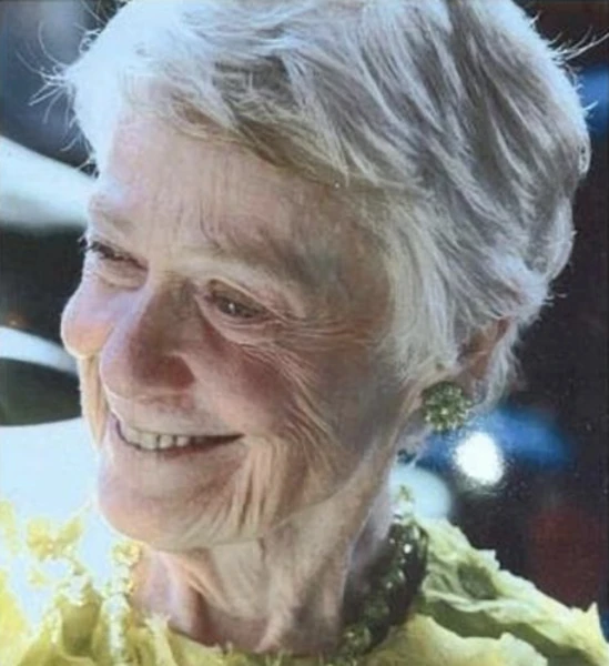 Smiling elderly woman with white hair and green earrings wearing a yellow top