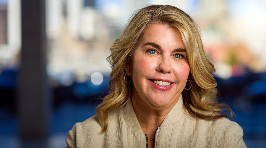 Smiling woman with blonde hair and blue eyes in a light jacket with a blurred city skyline behind her