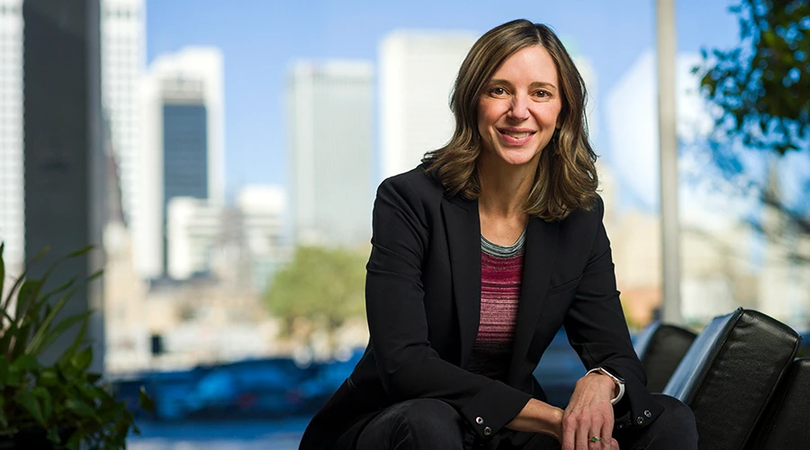 Woman in black blazer smiling with Tulsa city skyline visible through window behind her