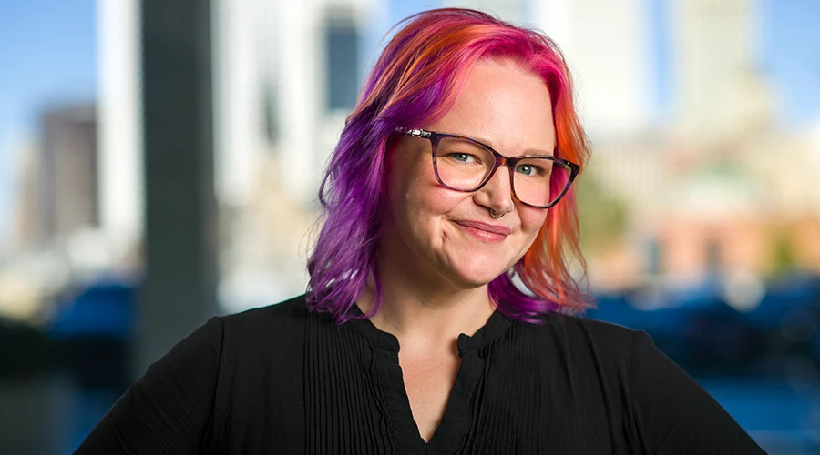 Smiling woman with colorful pink and orange hair and tortoiseshell glasses posed against a blurred Tulsa cityscape