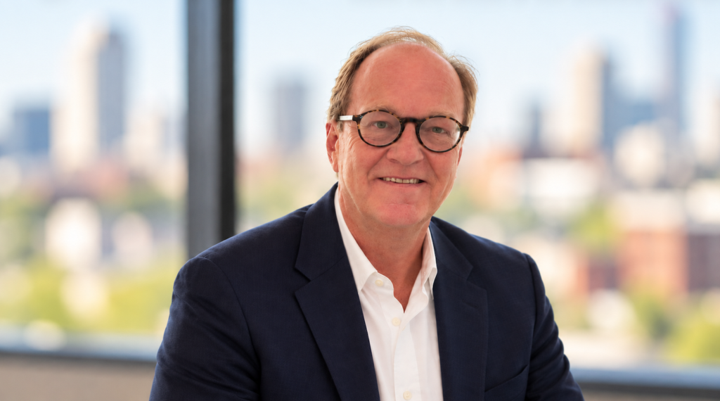 Smiling man in navy blazer and round glasses with Tulsa city skyline visible through window behind him